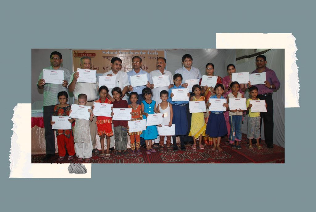 A group of children pose with school vouchers. Behind them, a group of adult women and men are also seen posing with the vouchers.