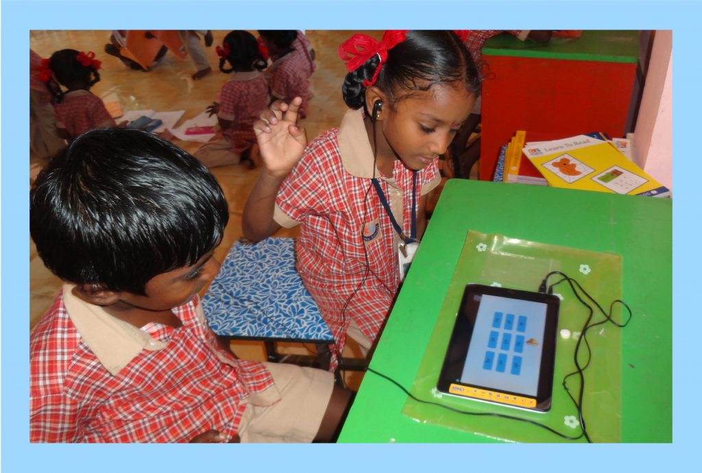 A photo of two young children, in their red and beige school uniforms, are seen sitting in front of a table with a learning tablet. The child on the left is seen wearing earphones that connect to the tablet she is learning from. The photo is placed against a light blue backdrop.