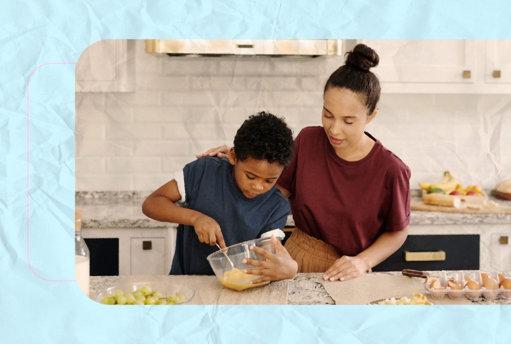 A photo of a young boy donned in a dark blue shirt whisking eggs in a bowl while his mother, wearing a maroon t-shirt, is supervising him with her arm around his shoulder. 