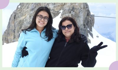 A mother and daughter, the former dressed in black sunglasses and black woollens and the latter dressed in light blue wolollens — pose for a photo in front of an ice hillock.