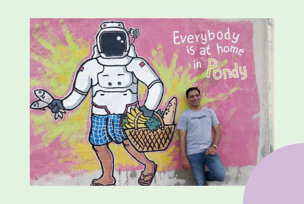 A middle-aged man wearing a grey t-shirt and dark blue jeans poses against a graffitied wall. The wall is painted in pink with a loud splash of yellow. In the yellow spot, is a mural of an astronaut wearing a mundu and carrying a basket of groceries and fish. Next to this, the wall reads 'Everybody is at home in Pondy'