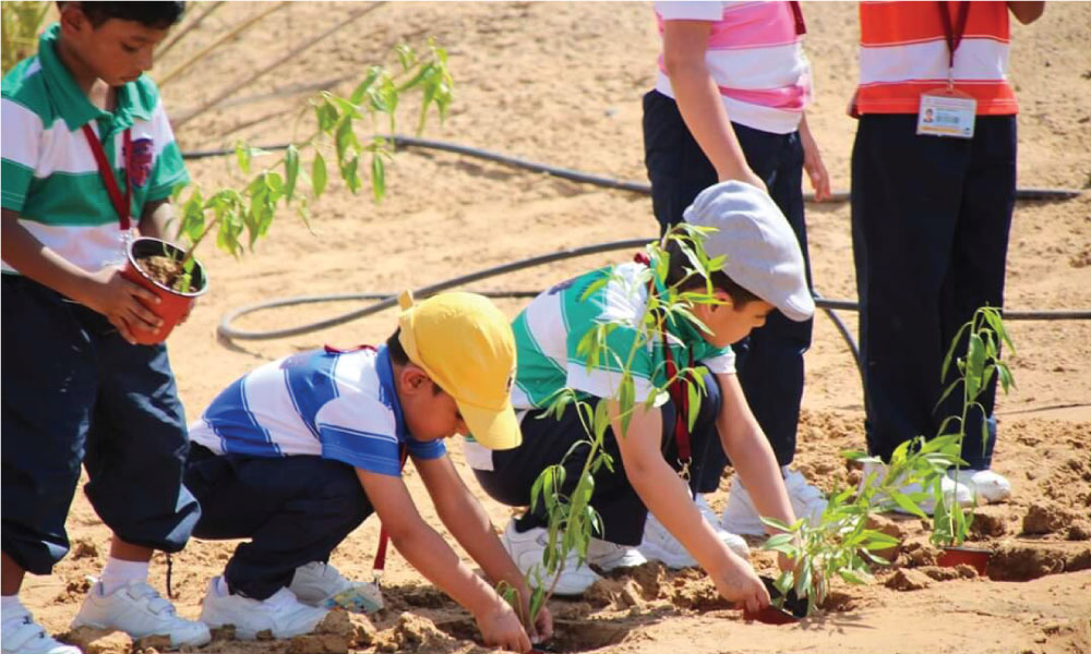 In this picture, we see five children dressed in striped shirts. Two of them are bent over and reaching for a plant sapling, presumably planting them in the ground. In the background, the other children standing..