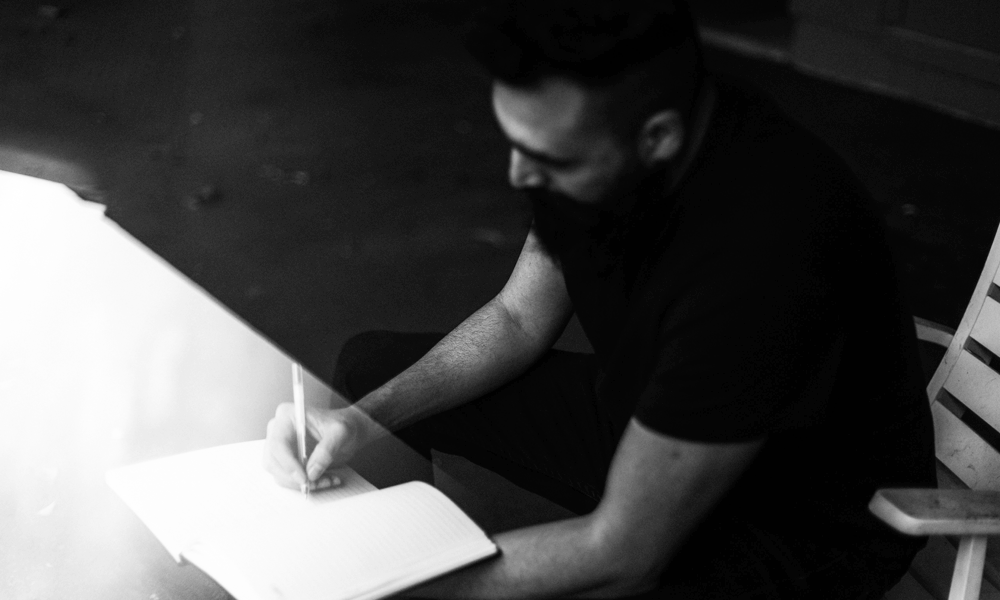 A black and white image of a bearded man writing in a diary.